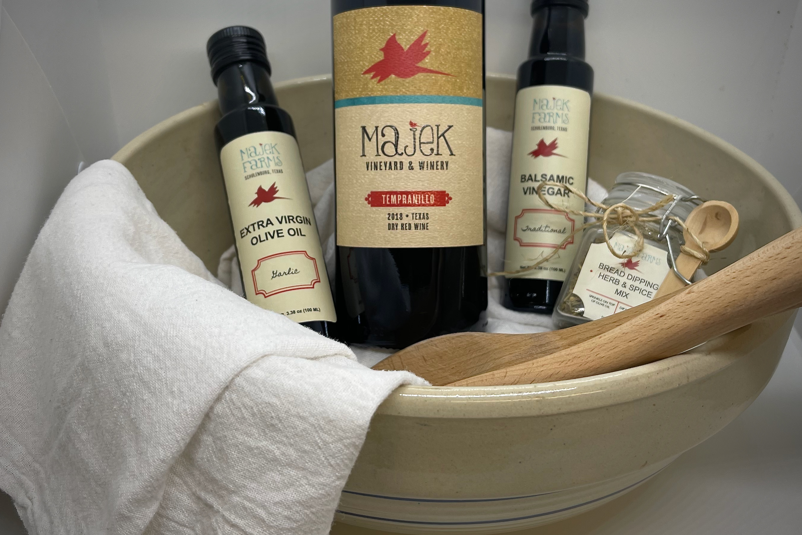 Mini bottles of olive oil, balsamic vinegar, a jar of dipping spice, and a bottle of red wine inside a big bowl with a white cloth.